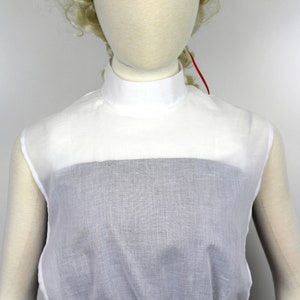 May include: White cotton mock neck shirt with a gathered bottom. The shirt is sleeveless and has a high neckline.