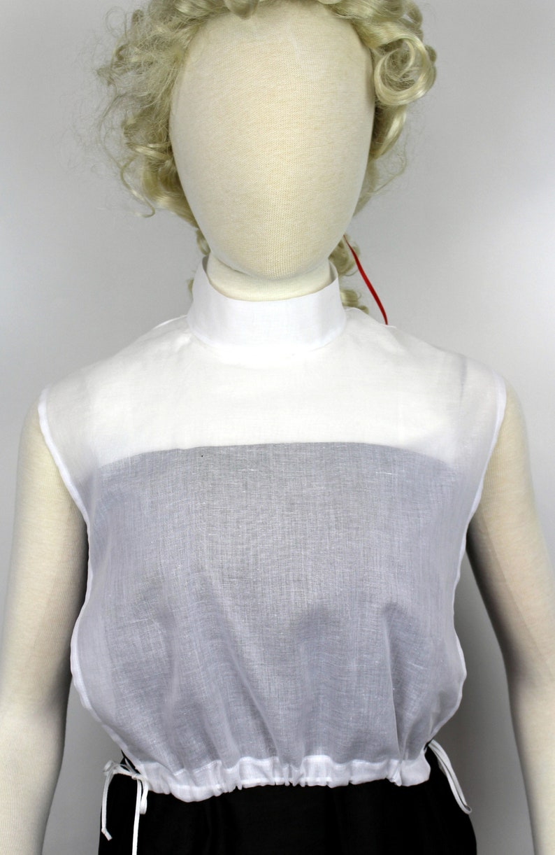 May include: White cotton mock neck shirt with a gathered bottom. The shirt is sleeveless and has a high neckline.