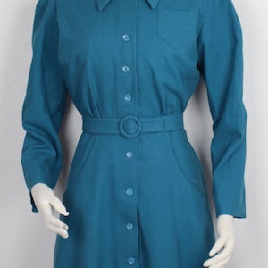 May include: A teal blue dress with a collared neckline, long sleeves, and a button-up front. The dress has a belt with a metal buckle and a pocket on the left side. The dress is worn by a mannequin with blonde hair and a teal blue hat.