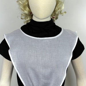 May include: White cotton bib with a gathered neckline and ties. The bib is worn over a black mock neck shirt.