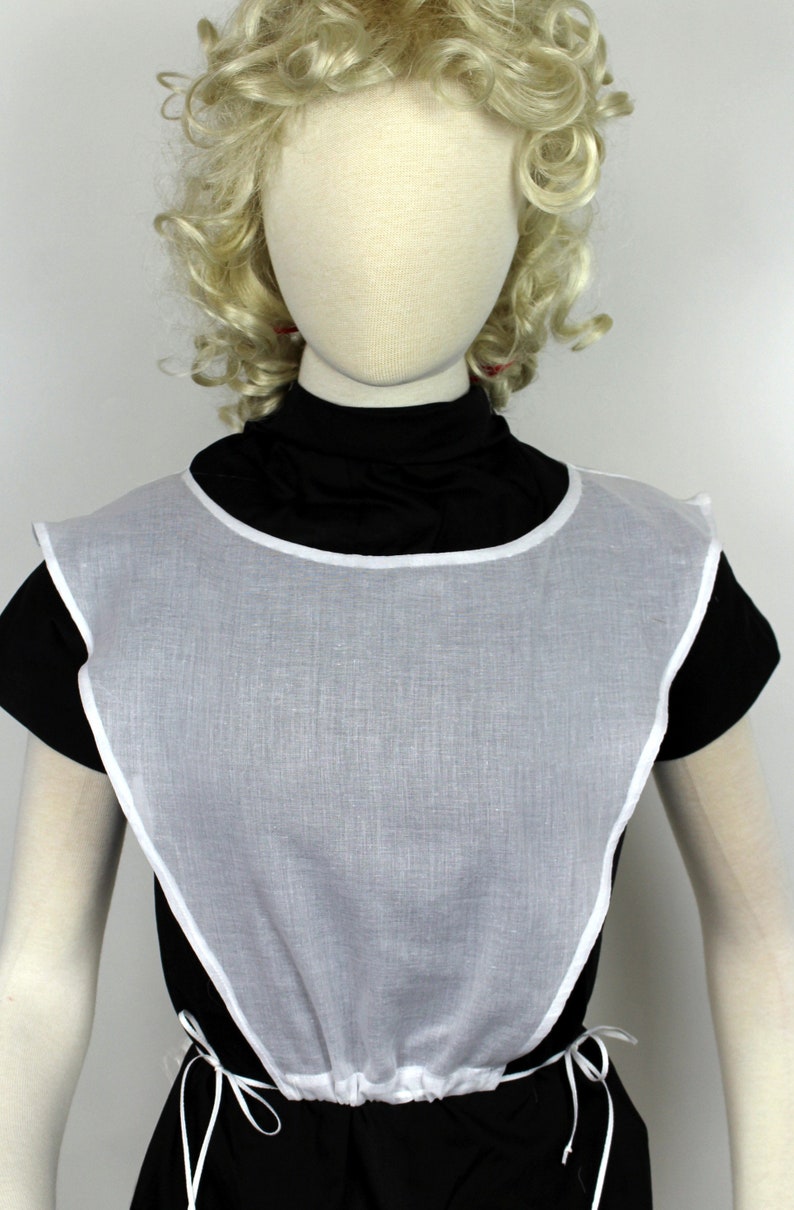 May include: White cotton bib with a gathered neckline and ties. The bib is worn over a black mock neck shirt.