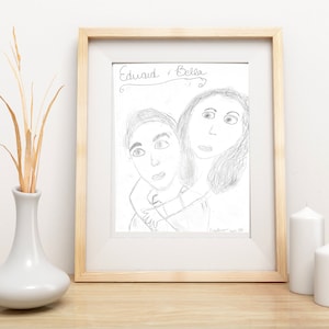 May include: A framed pencil sketch of two people, labeled "Edward & Bella". The artwork is in a light wood frame, displayed on a wooden surface with a white vase holding dried plants and two white candles.