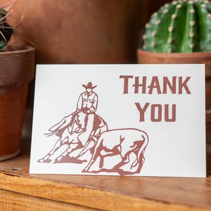 May include: A white card with a brown silhouette of a cowboy riding a horse and a cow. The card says "THANK YOU" in brown text.