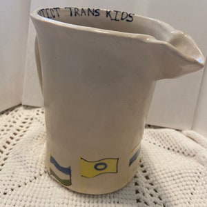May include: White ceramic pitcher with a blue and yellow flag design and the text "Protect Trans Kids" on the rim.