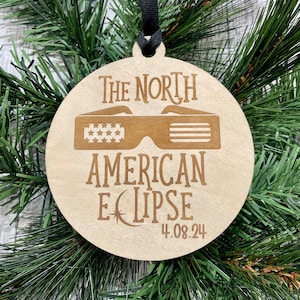 May include: A circular wooden ornament featuring the text "The North American Eclipse 4.08.24" and an illustration of eclipse glasses. It is suspended by a black ribbon and set against a backdrop of green pine needles.