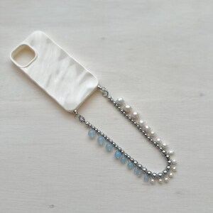 May include: Cream-colored phone case with a pearlescent finish, connected to a beaded strap. The strap features silver beads, freshwater pearls, and small blue teardrop-shaped beads. The case has a cutout for the camera.