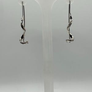 May include: A pair of silver-toned earrings. Each earring features a twisted, ribbon-like design suspended from a delicate chain. The earrings are displayed against a neutral background, highlighting their elegant, minimalist style. The earrings are a perfect accessory for any occasion.