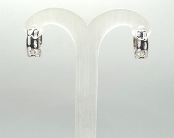 May include: A pair of silver-toned hoop earrings with a geometric design. The earrings are displayed on a white earring stand against a white background. The earrings have a rectangular pattern.
