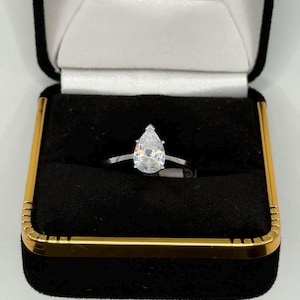 May include: A silver ring with a pear-shaped, clear gemstone, presented in a black jewelry box with a gold-colored trim. The ring's band is thin and delicate, showcasing the prominent gemstone.