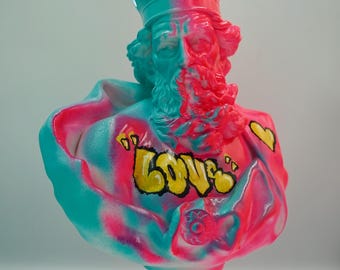 May include: A bust sculpture of a crowned figure, painted in vibrant teal and pink hues. The word "LOVE" is spray-painted in yellow on the figure's chest. The sculpture has a pedestal base and is likely a decorative art piece.