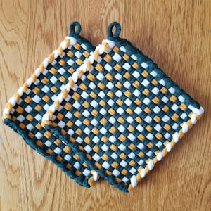 Op de afbeelding: Twee geweven pannenlappen met een groen, wit en goud geruit patroon. Elke pannenlap heeft een lus om op te hangen en een groene rand. De pannenlappen zijn gemaakt van geweven materiaal.