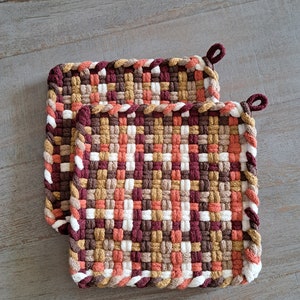 May include: Two square woven pot holders in shades of brown, white, and peach. The pot holders have a looped corner for hanging. The weave pattern is tight, and the edges are finished with a braided border.