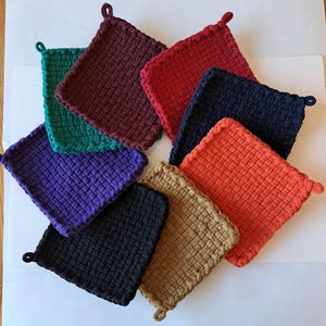 Peut inclure: Huit maniques tissées de différentes couleurs, dont le rouge, l'orange, le violet, le vert, le marron, le noir et le beige. Les maniques sont disposées en cercle sur une surface blanche.