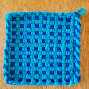 May include: A blue and green woven pot holder with a knotted loop handle. The pot holder is made of cotton yarn and has a checkered pattern.