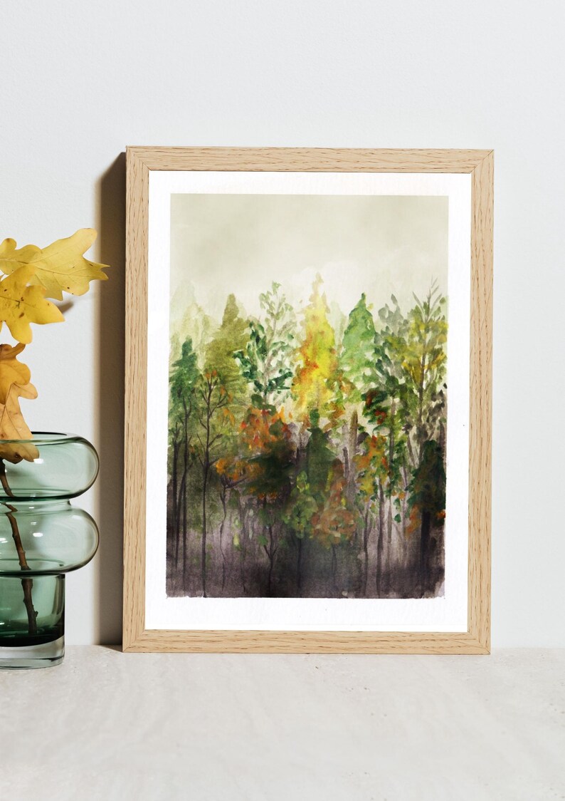 May include: A framed watercolor painting depicting a dense forest scene with varying shades of green, yellow, and brown foliage. The artwork features tall, slender tree trunks and a soft, hazy background, all encased in a light-toned wooden frame.