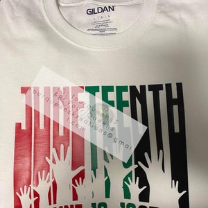 May include: White t-shirt with a graphic design featuring the word "THIRTEENTH" in red, green, and black, with raised hands and the date "JUNE 19, 1865". The shirt has a crew neck and the Gildan brand label.