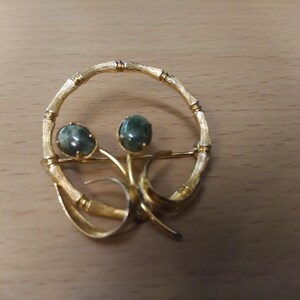 May include: A gold-toned brooch with a circular design featuring two green cabochon stones. The brooch has a delicate, intricate design with a stylized floral motif.