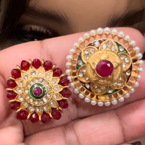 May include: Two gold-toned, round, decorative jewellery pieces with red and white stones. The larger piece has a pearl border and a red stone in the centre. The smaller piece has a red stone border and a smaller red stone in the centre.