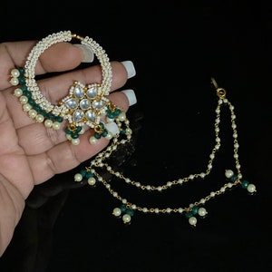 May include: A gold nose ring with a white pearl and green bead chain. The ring has a large, ornate flower design with clear stones.