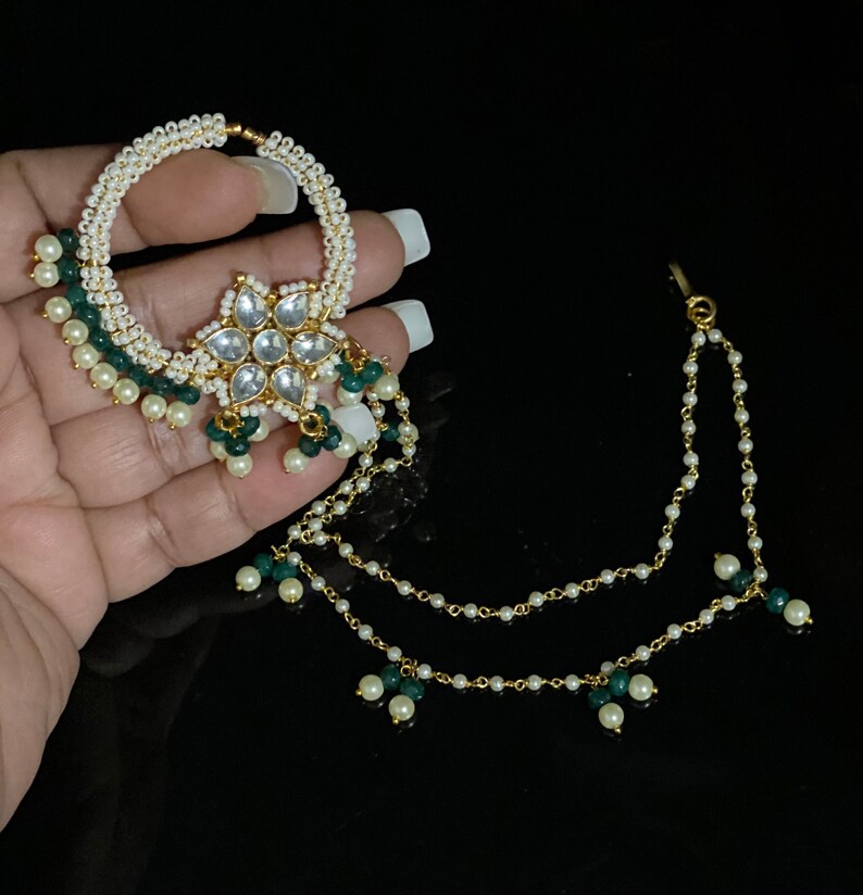 May include: A gold nose ring with a white pearl and green bead chain. The ring has a large, ornate flower design with clear stones.