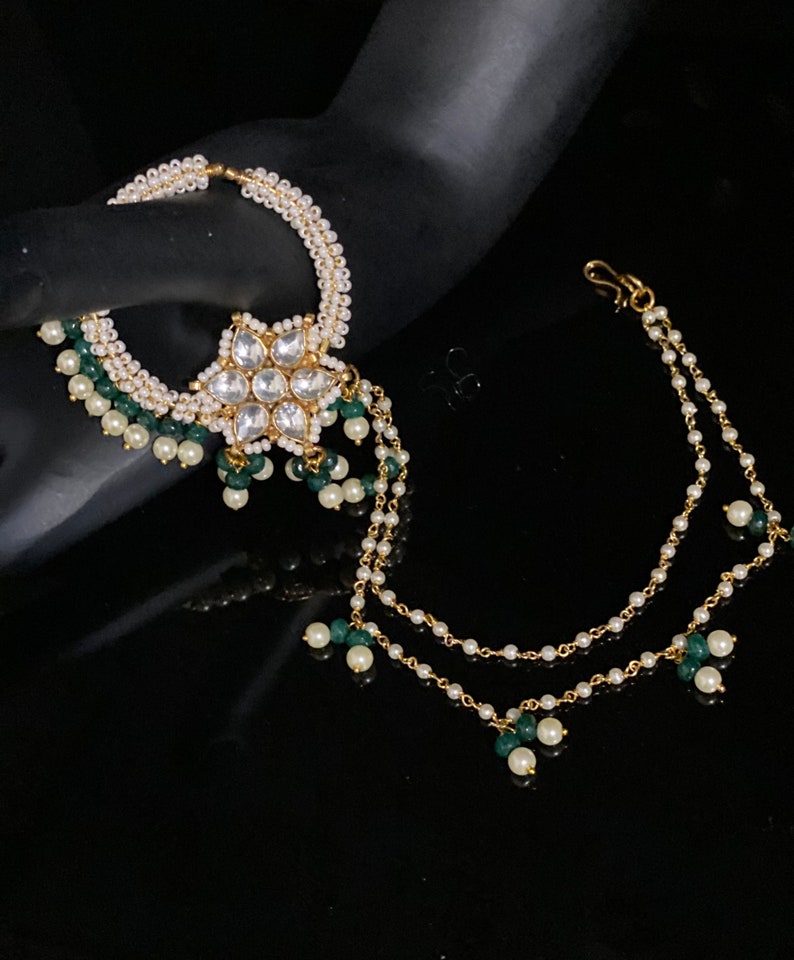 May include: A gold nose ring with a white flower design and green and white beads. The ring is attached to a chain with green and white beads.