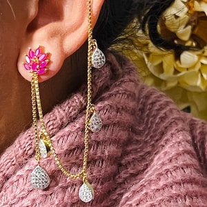 May include: Gold ear cuff with a dangling chain and pink and clear crystal teardrop charms. The ear cuff is worn on the upper part of the ear.