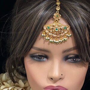 May include: A gold-tone headpiece with green and white stones. The headpiece is designed to be worn on the forehead and has a teardrop shape.