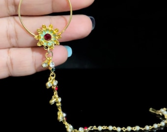 May include: Gold nose ring with a chain and two flower-shaped charms. The flowers are red, green, and white.
