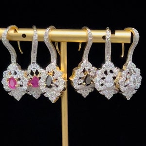 May include: A set of four dangle earrings with a floral design. Two earrings feature a red gemstone, and two earrings feature a black gemstone. The earrings are on a gold stand against a black background.