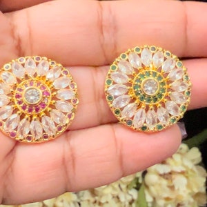 May include: A pair of gold-tone earrings with a circular design. The earrings are adorned with white and green stones, and the center of each earring features a larger white stone.