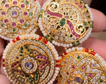 May include: Four gold-toned round ornaments with intricate designs featuring white, pink, and green gemstones. The ornaments are surrounded by a border of white pearls.