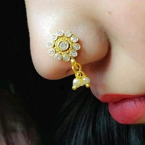 May include: A gold nose ring with a round, white stone center surrounded by smaller white stones. The ring has a chain with a pearl drop.
