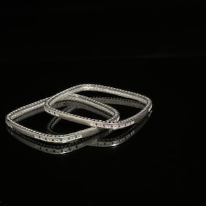 May include: Three silver square bangles with a row of small green and pink gemstones around the edge.