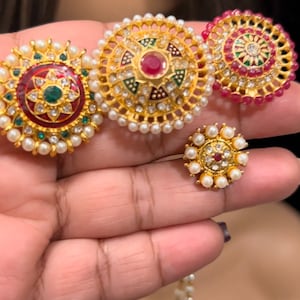 May include: Four gold-toned, round, decorative pieces with red, green, and white enamel and pearl accents. The pieces are arranged in a row on a hand.