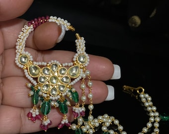 May include: A gold nose ring with a large, ornate design featuring white and green gemstones. The ring is attached to a chain with white pearls and green and red gemstones.