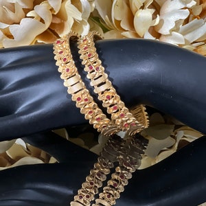 Op de afbeelding: Twee gouden armbanden met rode stenen accenten. De armbanden zijn ingewikkeld ontworpen met bloemmotieven.