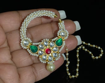 May include: A gold nose ring with a pearl chain. The ring is decorated with white pearls, red and green gemstones, and a large pearl drop.