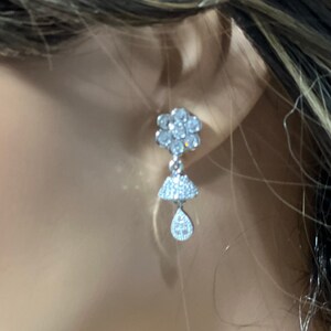 Puede incluir: Pendientes de plata con forma de flor en la parte superior y forma de lágrima en la parte inferior, ambos adornados con piedras de circonita cúbica transparentes.