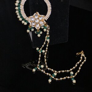 May include: A gold nose ring with a white pearl and green bead chain. The ring has a large, clear, flower-shaped stone in the center.