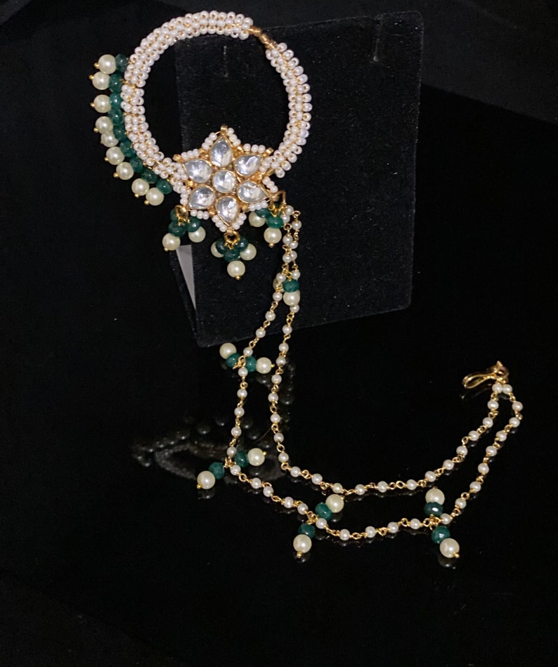May include: A gold nose ring with a white pearl and green bead chain. The ring has a large, clear, flower-shaped stone in the center.