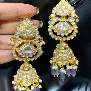 May include: A pair of gold earrings with intricate designs and white stones. The earrings have a dangling bell shape with purple beads.