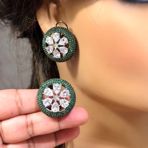 Può includere: Un paio di orecchini rotondi con un designo verde e bianco. Gli orecchini presentano un gruppo di pietre chiare a forma di pera circondate da pietre verdi più piccole.