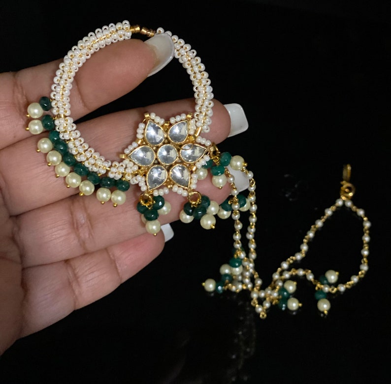 May include: A gold nose ring with a star-shaped pendant adorned with white stones and green beads. The ring is attached to a chain with white and green beads.