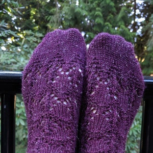 May include: A pair of purple knitted socks with a delicate openwork pattern. The socks have a ribbed cuff and a sparkly, textured appearance. The socks are resting on a black railing with a blurred green background.