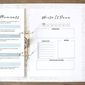 May include: A white binder with two pages open. The left page has the title "Morning Moments" and lists five steps for a morning routine: silence, affirmations, visualize, gratitude, and journal/move. The right page has the title "Write It Down" and includes sections for goals, gratitude, and what you hope for today.