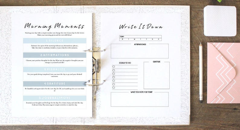 May include: A white binder with two pages open. The left page has the title "Morning Moments" and lists five steps for a morning routine: silence, affirmations, visualize, gratitude, and journal/move. The right page has the title "Write It Down" and includes sections for goals, gratitude, and what you hope for today.