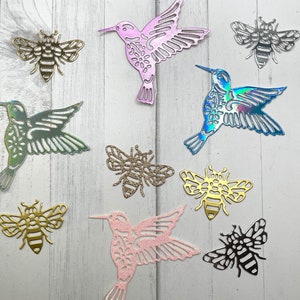 May include: Assortment of die-cut metal hummingbirds and bees in various colors. The hummingbirds are pink, green, and blue with intricate wing and body details. The bees are gold, silver, and black, some with glitter accents. The shapes are scattered on a light wood grain surface.