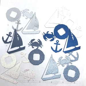 May include: A collection of blue, silver, and white paper cutouts of nautical themed shapes, including sailboats, anchors, crabs, and life preservers.