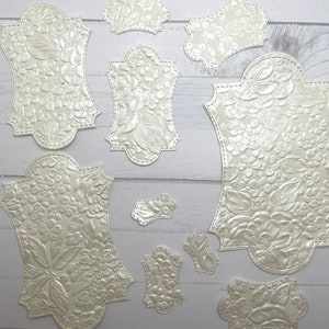 May include: A set of ten white, embossed paper labels with a floral design. The labels are in various sizes and shapes, with scalloped edges.