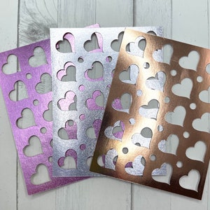 May include: Three sheets of decorative paper in shades of purple, silver, and copper. Each sheet features a pattern of heart-shaped cutouts and small circular perforations. The paper has a metallic sheen, creating a shimmering effect.